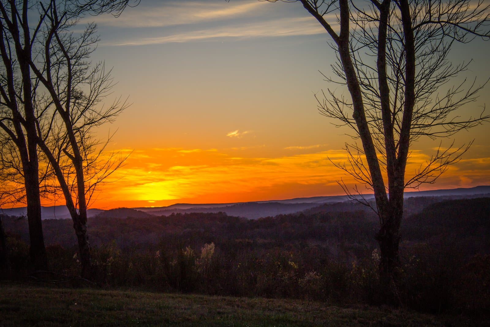 The sun sets in orange behind bare trees and rolling wooded ridges, seen from atop Taylor's Ridge in Chattooga County, Ga., Nov. 26, 2015. Illustrative image; it does not show the Burke family's home or the fire. Photo: Shane Clements, CC BY 2.0, via Wikimedia Commons. Source page: https://commons.wikimedia.org/wiki/File:Chattooga_County,_Georgia_-_Sunset.jpg