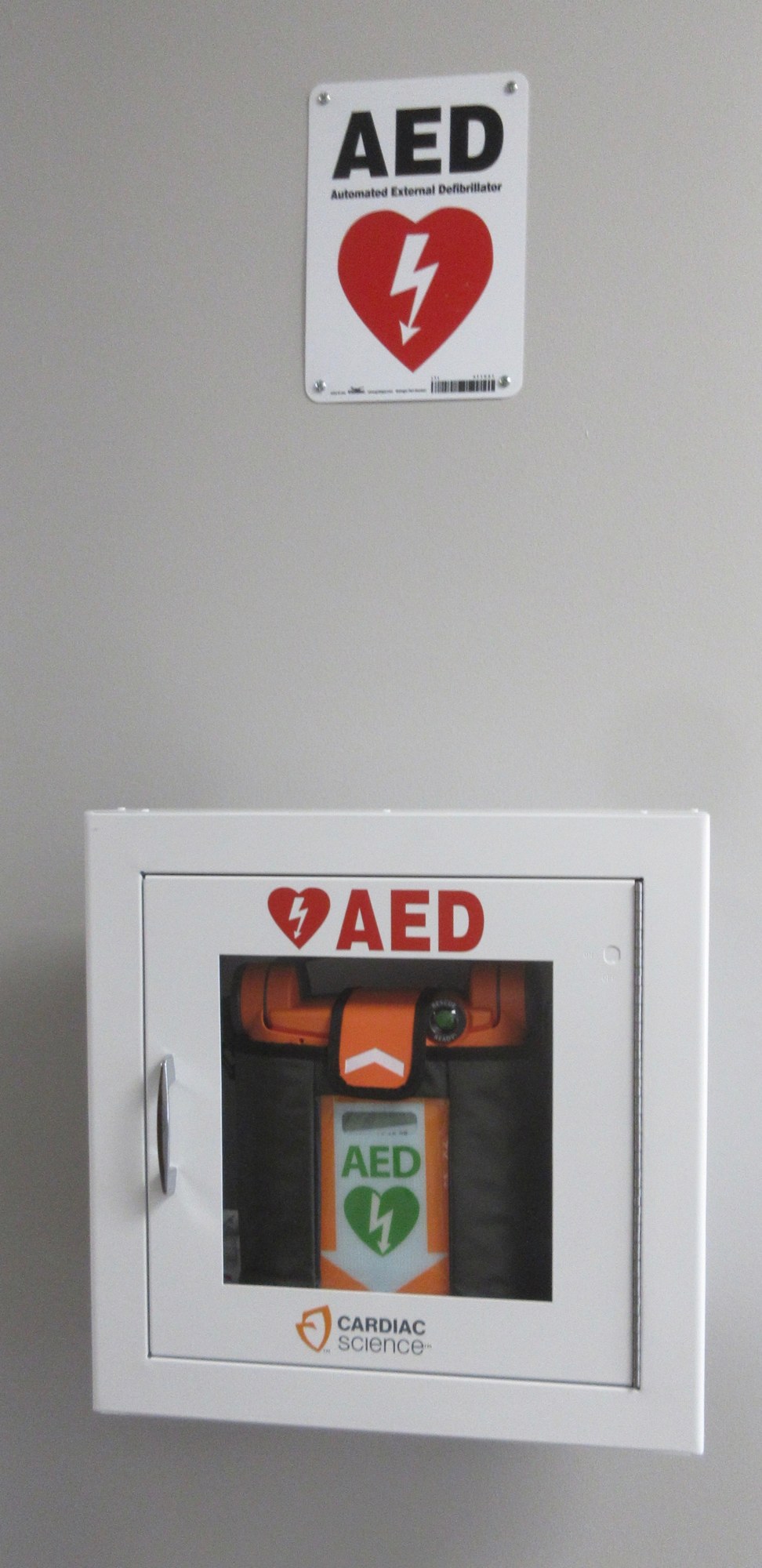 A wall-mounted white cabinet holding an orange automated external defibrillator, with a red heart AED sign mounted above it.
