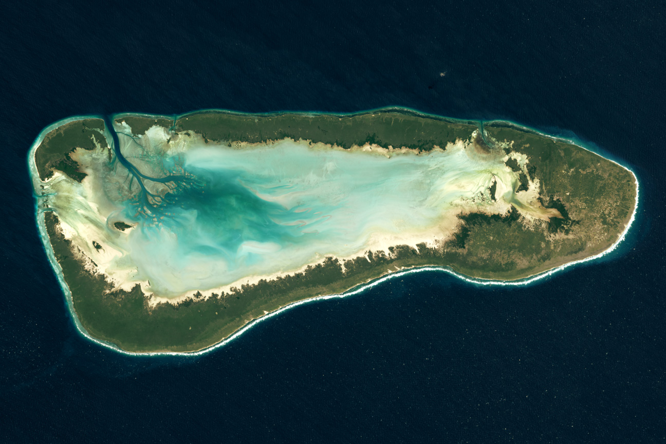 Satellite view of an elongated coral atoll ringed by green islands around a pale turquoise lagoon in dark blue ocean.