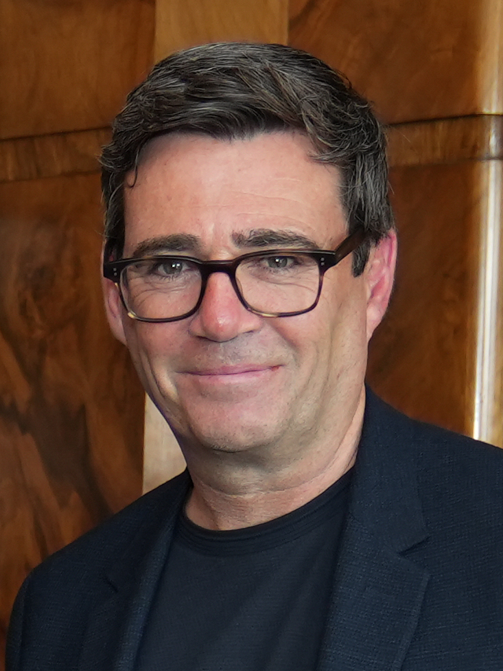A smiling man with dark hair and dark-rimmed glasses, in a dark jacket, photographed indoors against a wood-paneled background.