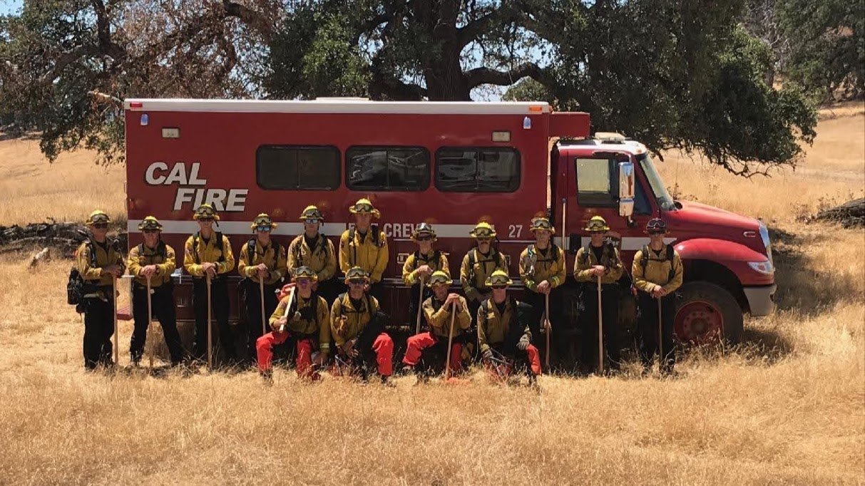 About twenty firefighters in yellow shirts and helmets, holding hand tools, pose in two rows in dry grass in front of a red CAL FIRE crew truck.