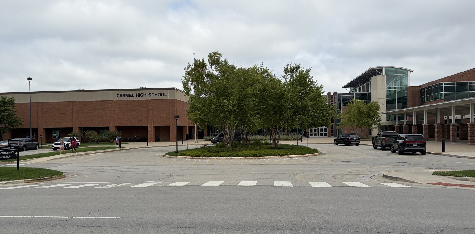 A long brick school building with a sign reading Carmel High School, a landscaped traffic circle and parked cars under an overcast sky.