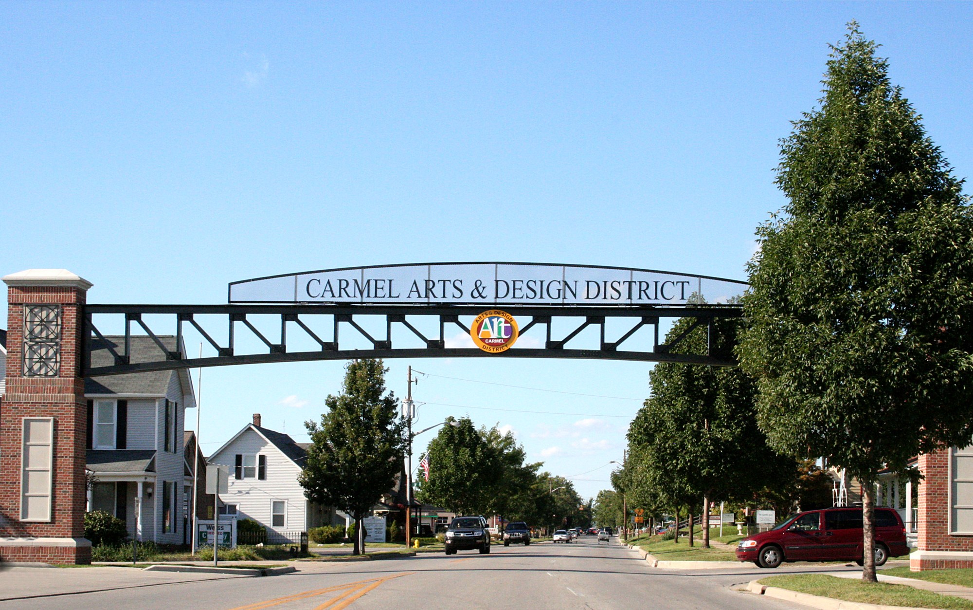 A steel archway reading Carmel Arts and Design District spans a street lined with trees and small houses under a clear blue sky.