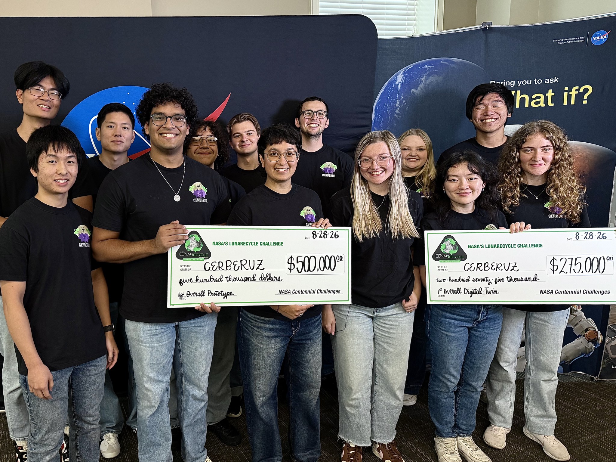 Fourteen students stand in front of NASA backdrops holding two large checks for $500,000 and $275,000.