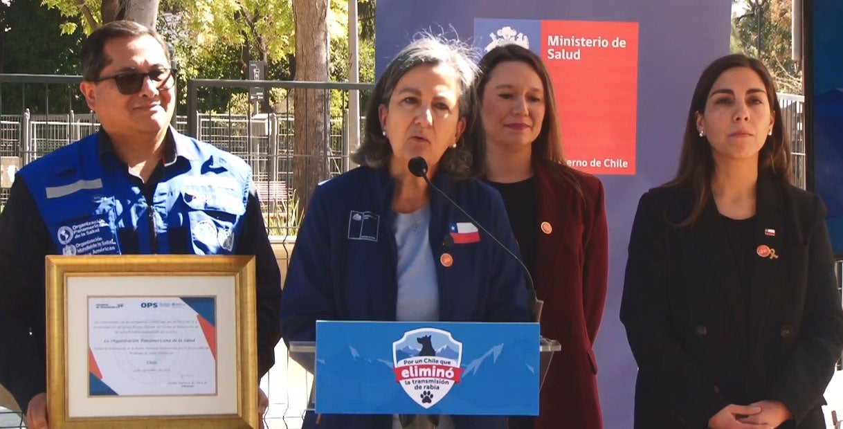 Chile's Minister of Health, Dr May Chomalí, speaks at an outdoor podium beside a PAHO/WHO representative holding a framed certificate, with Ministry of Health banners visible during the validation ceremony.