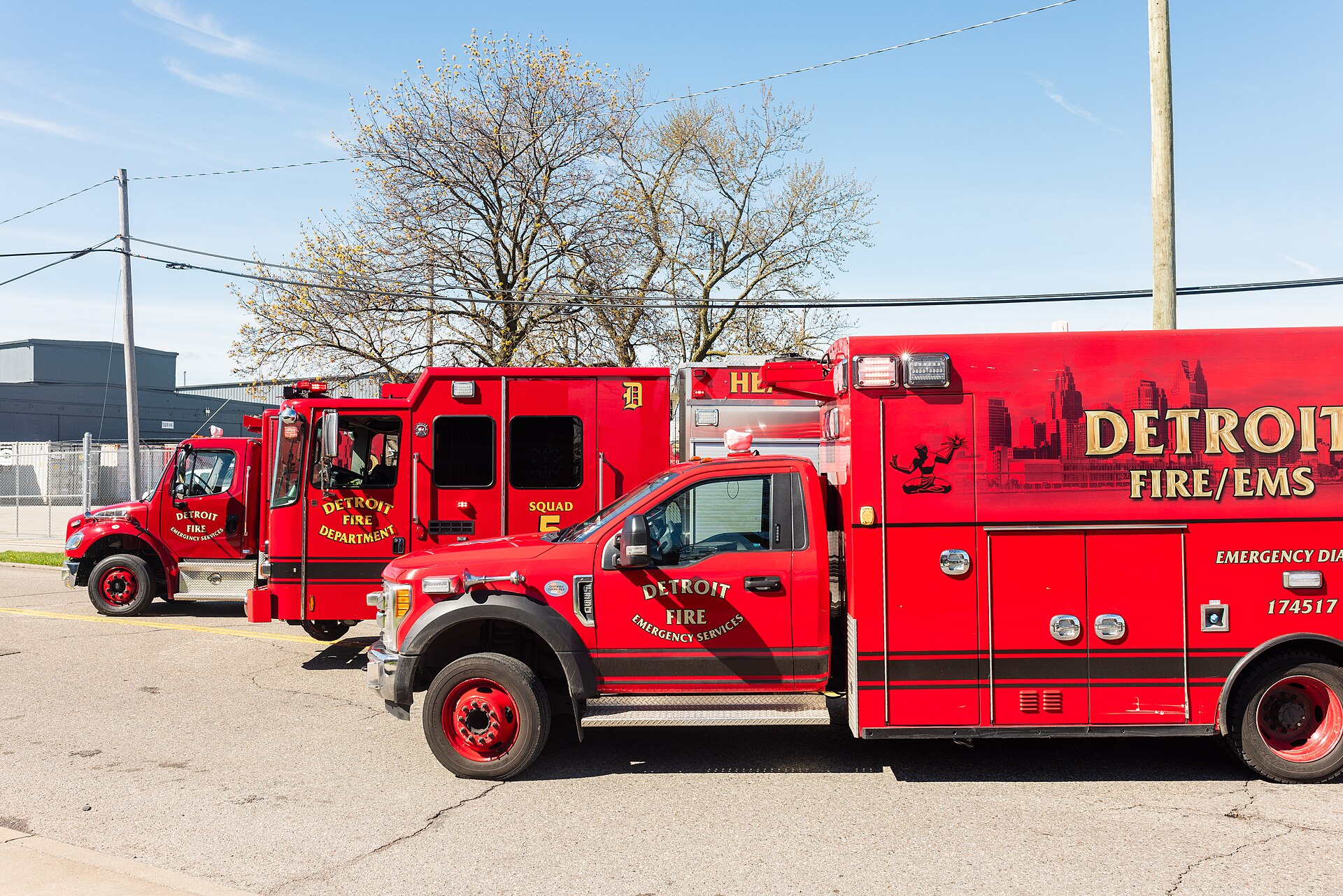 Red Detroit Fire/EMS ambulances and a fire vehicle parked on a paved lot under a blue sky with bare trees.