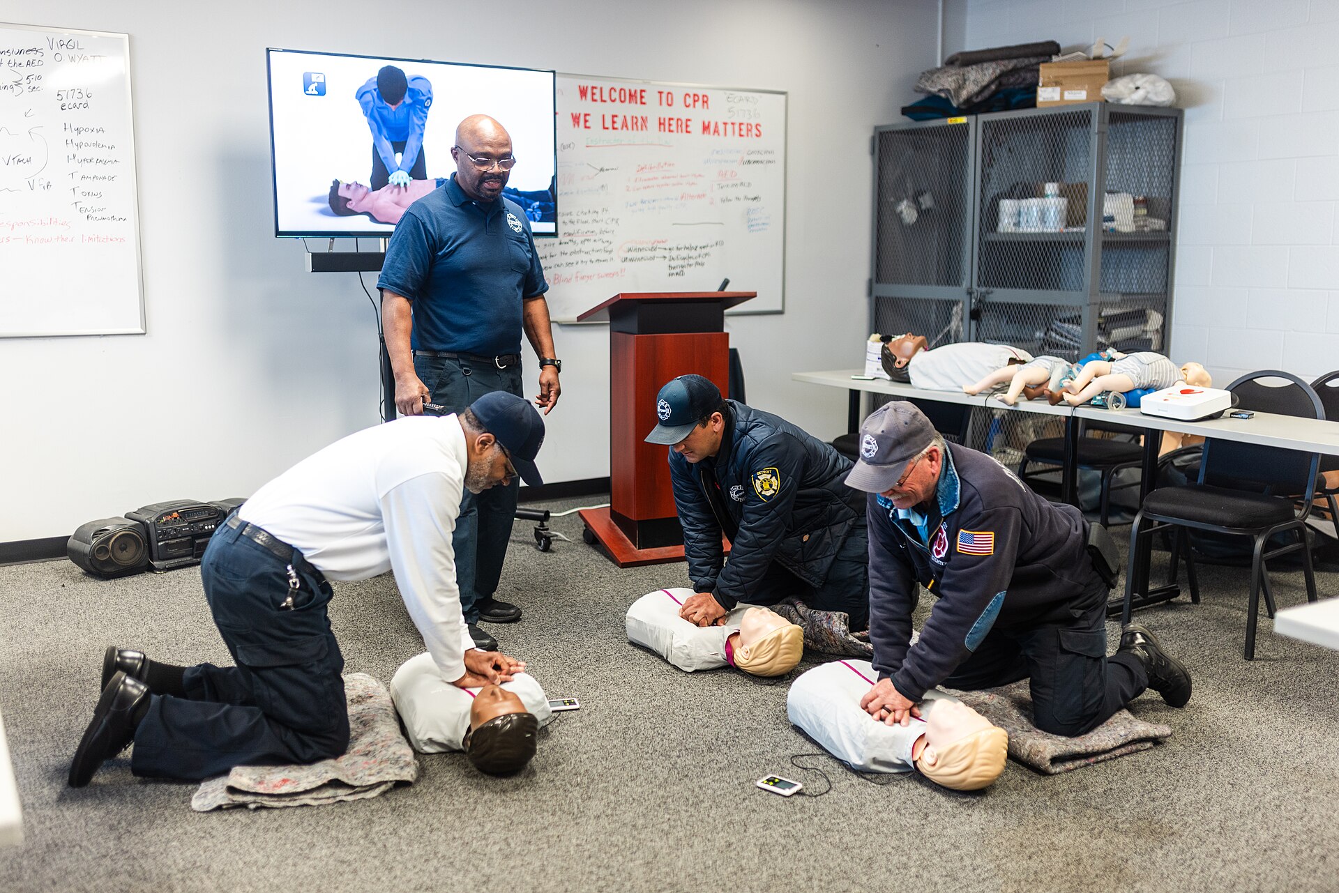 Three people kneel on a classroom floor practicing chest compressions on CPR training manikins as an instructor watches.