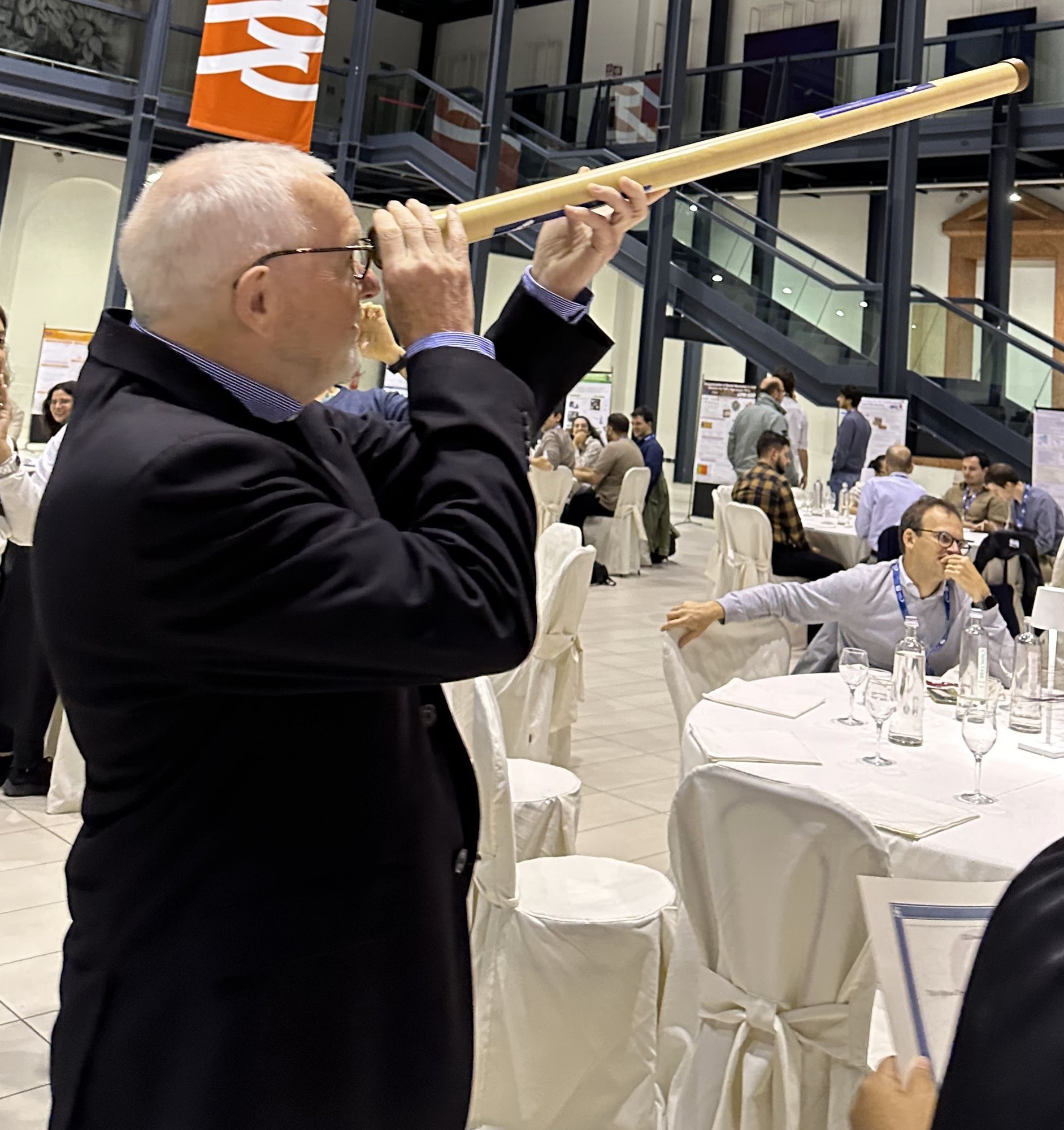 An older man with white hair and glasses, in a dark jacket, looks through a long wooden replica telescope inside a crowded conference hall.