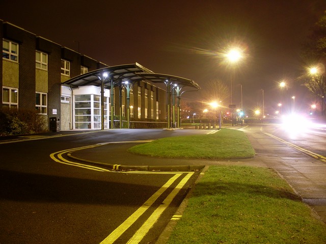 A lit, glass-fronted entrance canopy at the Freeman Hospital in Newcastle upon Tyne at night, with street lamps and yellow road markings in the foreground.
