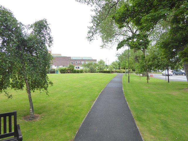 A paved footpath curving across a wide green lawn lined with trees, with hospital buildings in the distance and a bench in the foreground.