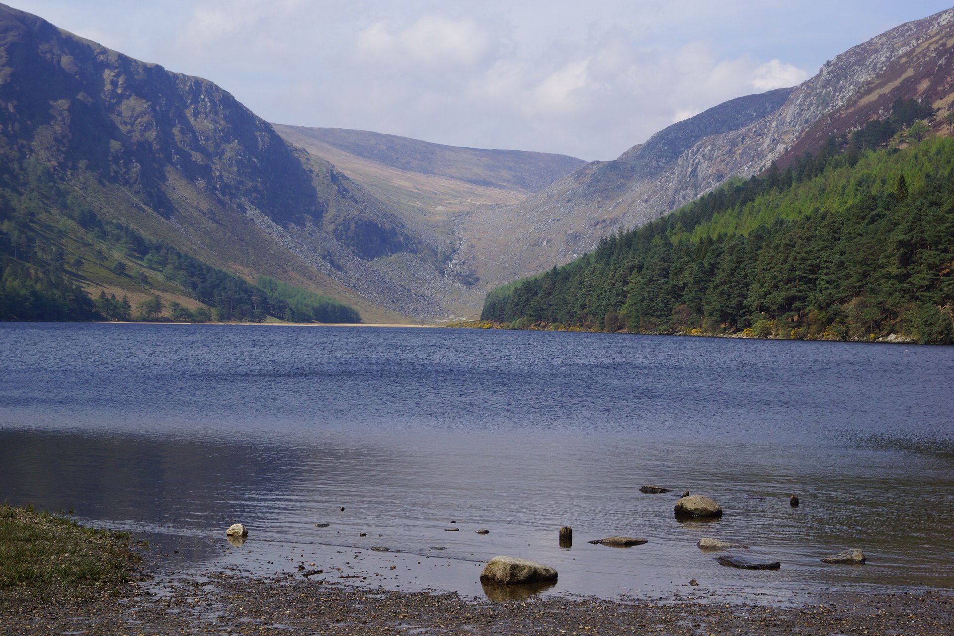 A calm lake with stones along its near shore lies at the foot of steep, partly wooded mountains under a pale cloudy sky.