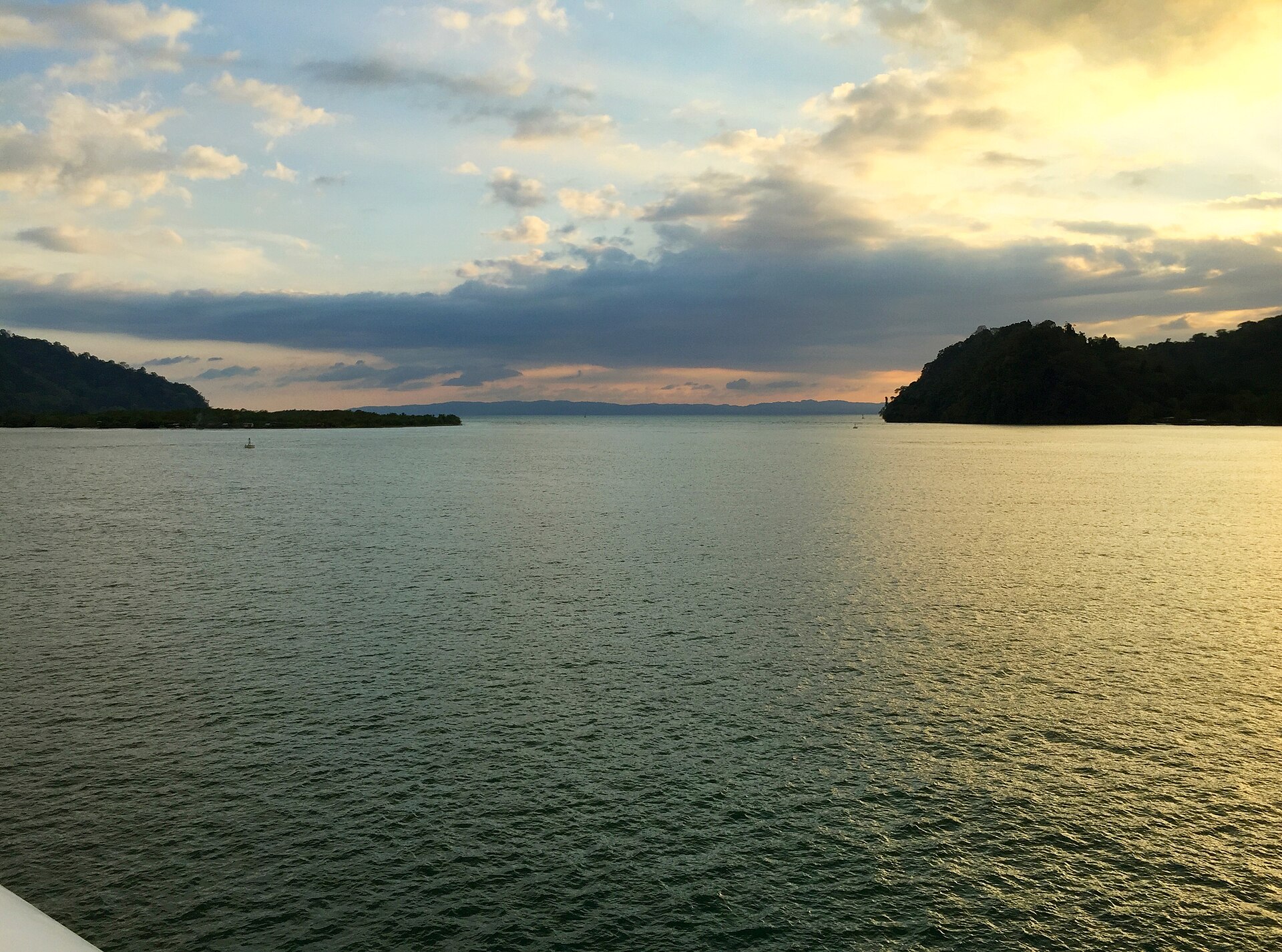 Calm water at sunset with forested headlands on the horizon under a partly cloudy sky.