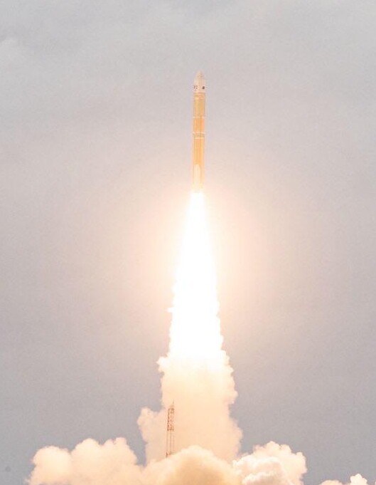 A tall white and orange rocket rising on a bright column of flame and smoke into an overcast sky.