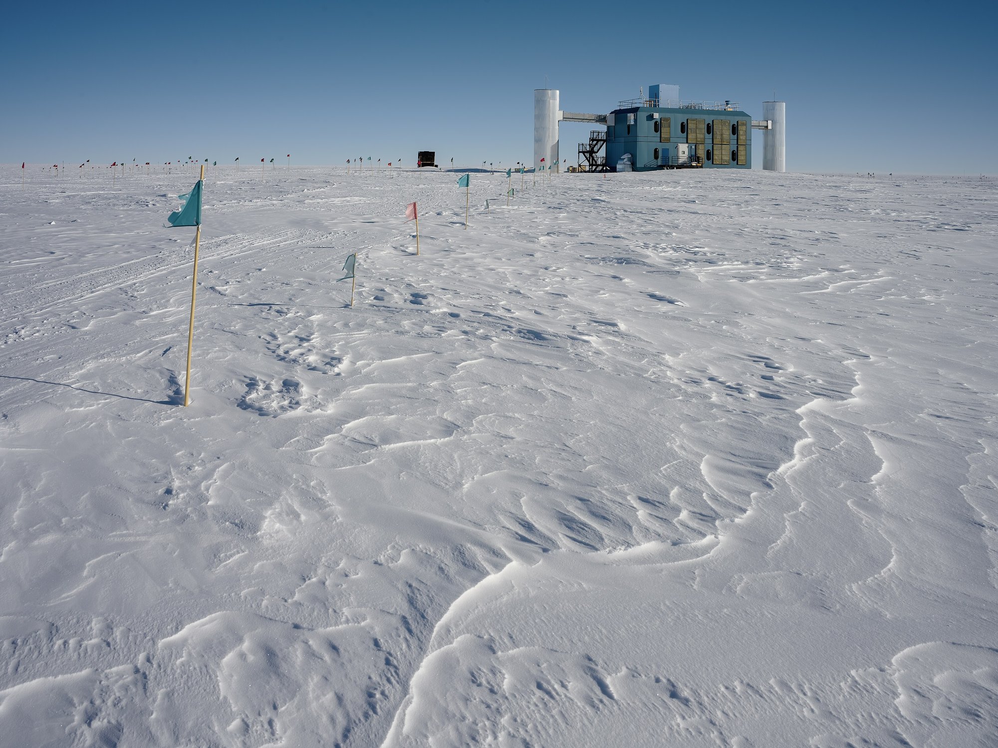 A blue and gold research building with two white towers stands on a vast, wind-scoured snowfield under a clear blue sky, with marker flags in the foreground.
