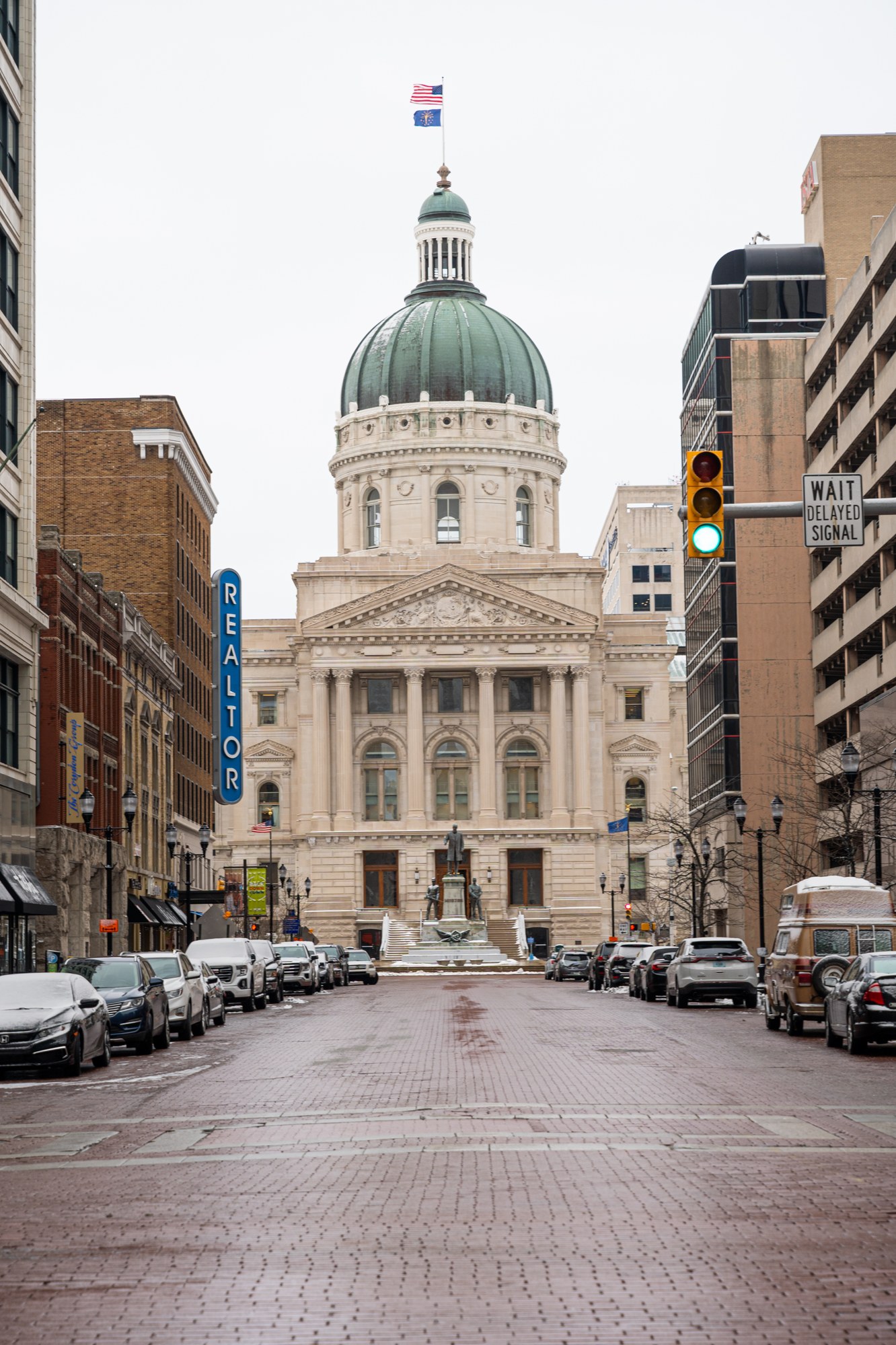The domed Indiana Statehouse at the end of a wet brick street lined with buildings and parked cars.