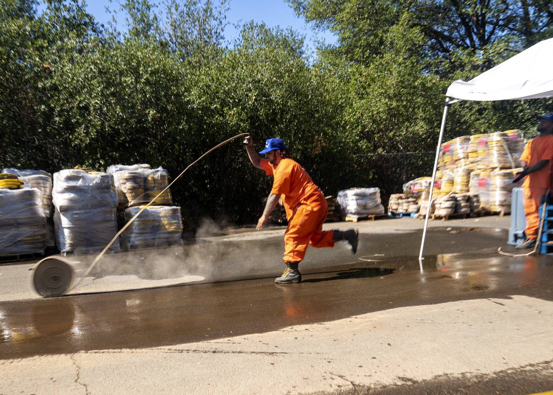 A man in an orange uniform and blue cap swings a long fire hose across wet pavement beside stacked pallets of supplies under a white canopy.
