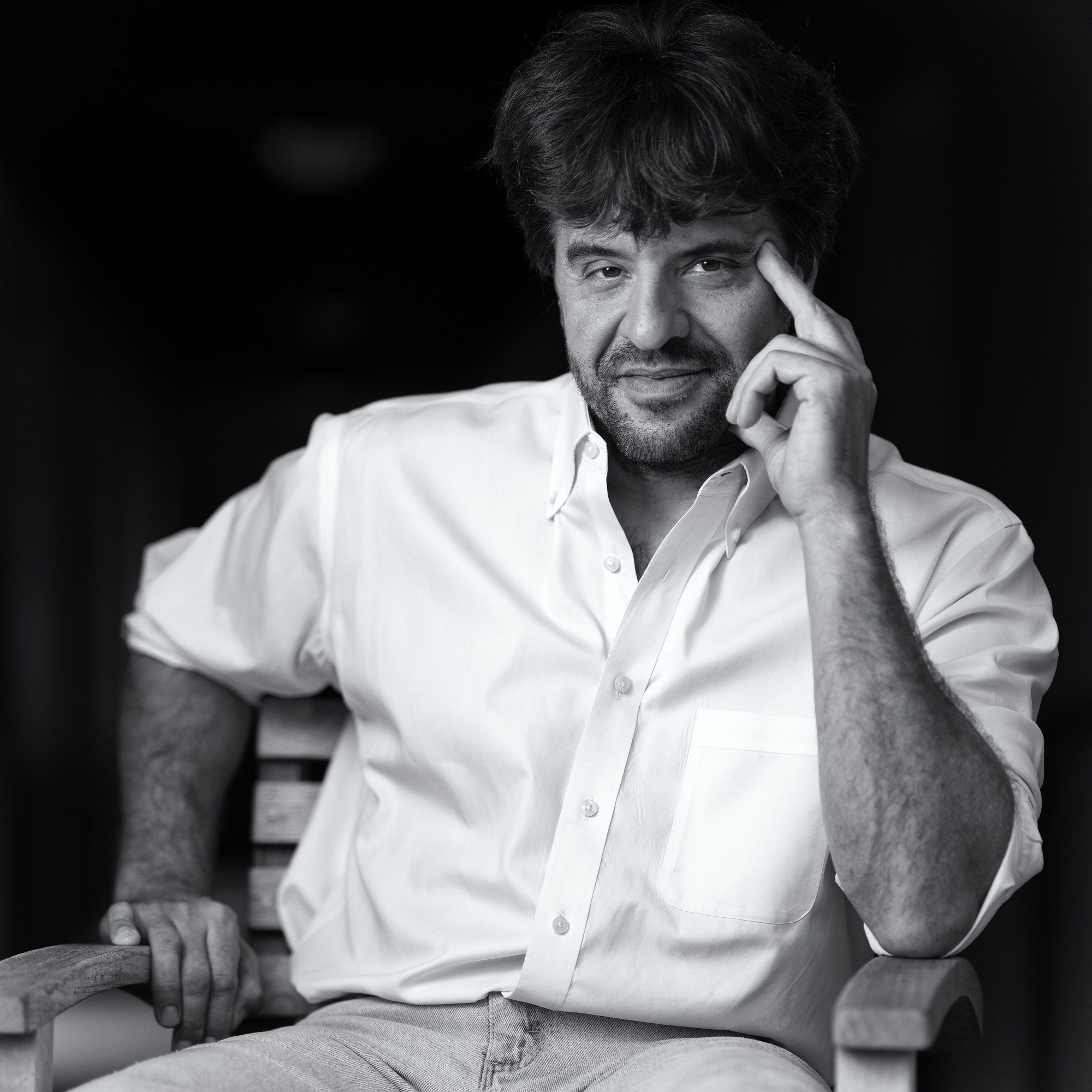 A black-and-white portrait of a man with dark, tousled hair in an open-collared white shirt, seated on a wooden bench with one hand raised to his temple.
