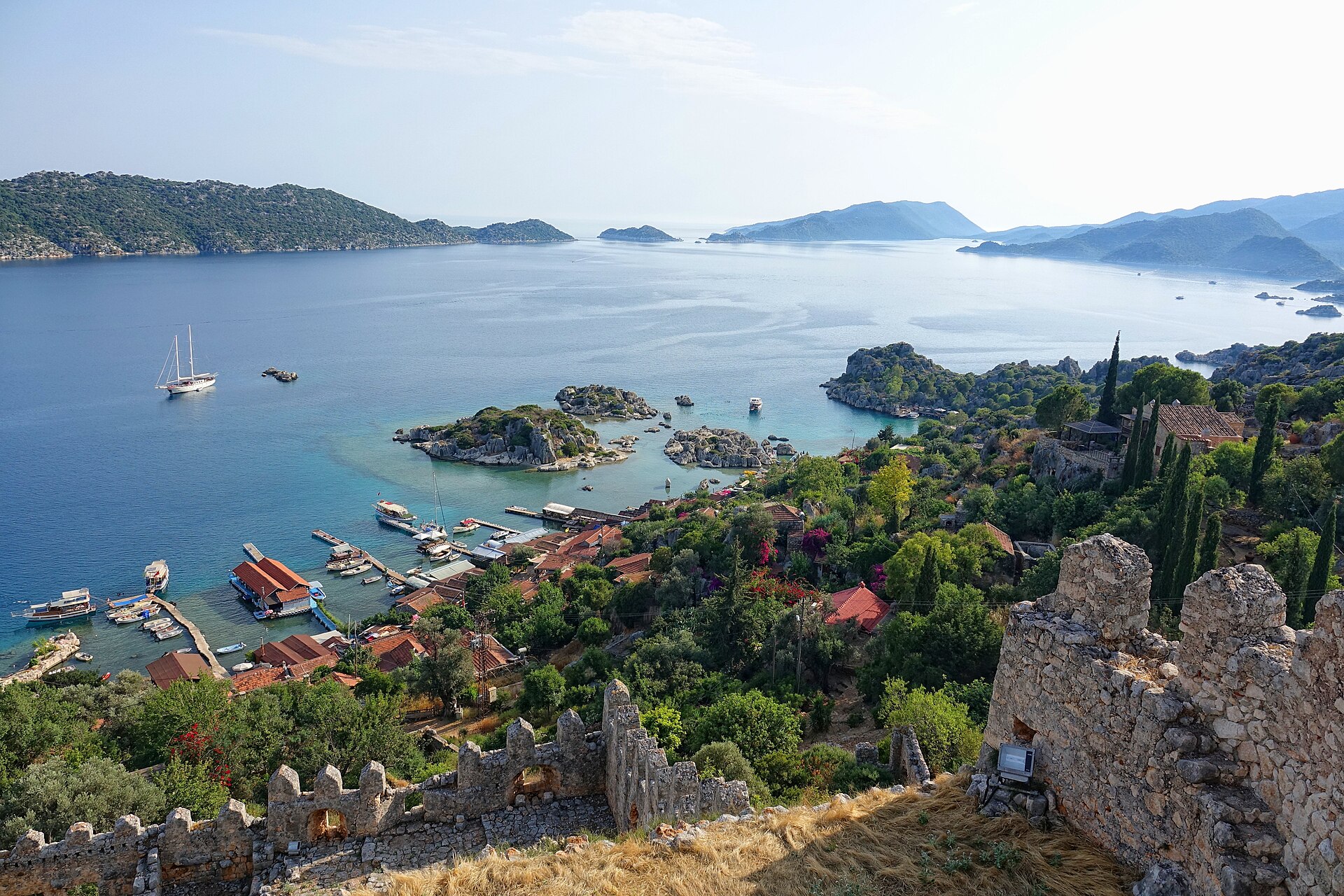View from a hilltop fortress over a village, a sheltered bay with boats and the long low profile of Kekova Island across blue water.