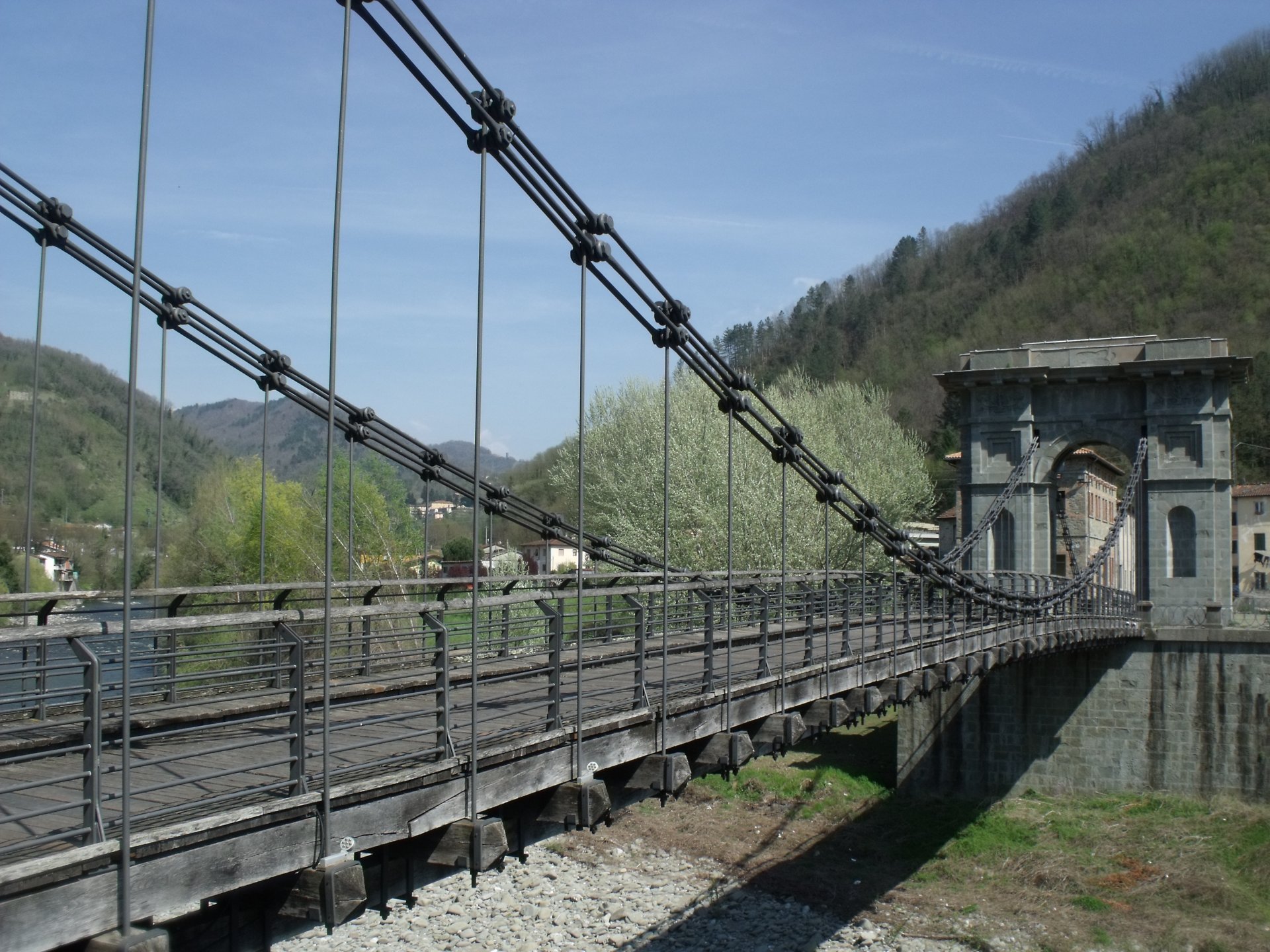 A long iron chain suspension bridge with a stone gateway at its far end spans a broad, stony riverbed between green wooded hills under a blue sky.