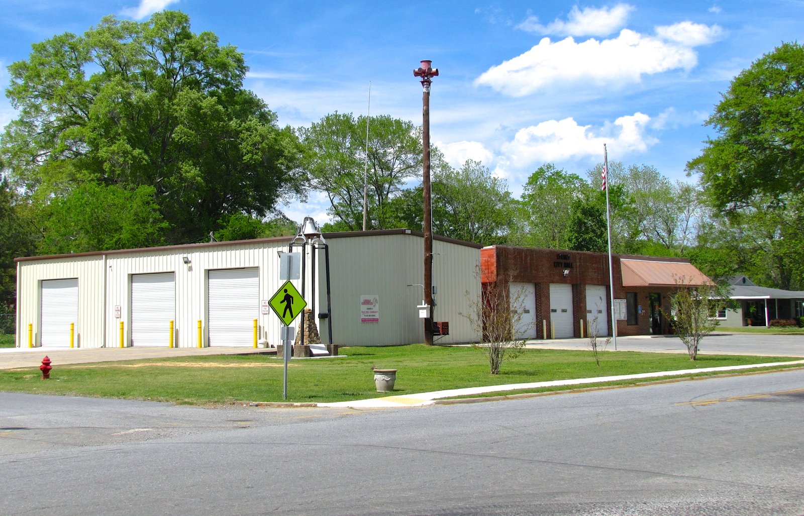 A white metal building with three roll-up bay doors stands beside a small brick city hall with an American flag, on a grassy corner lot.