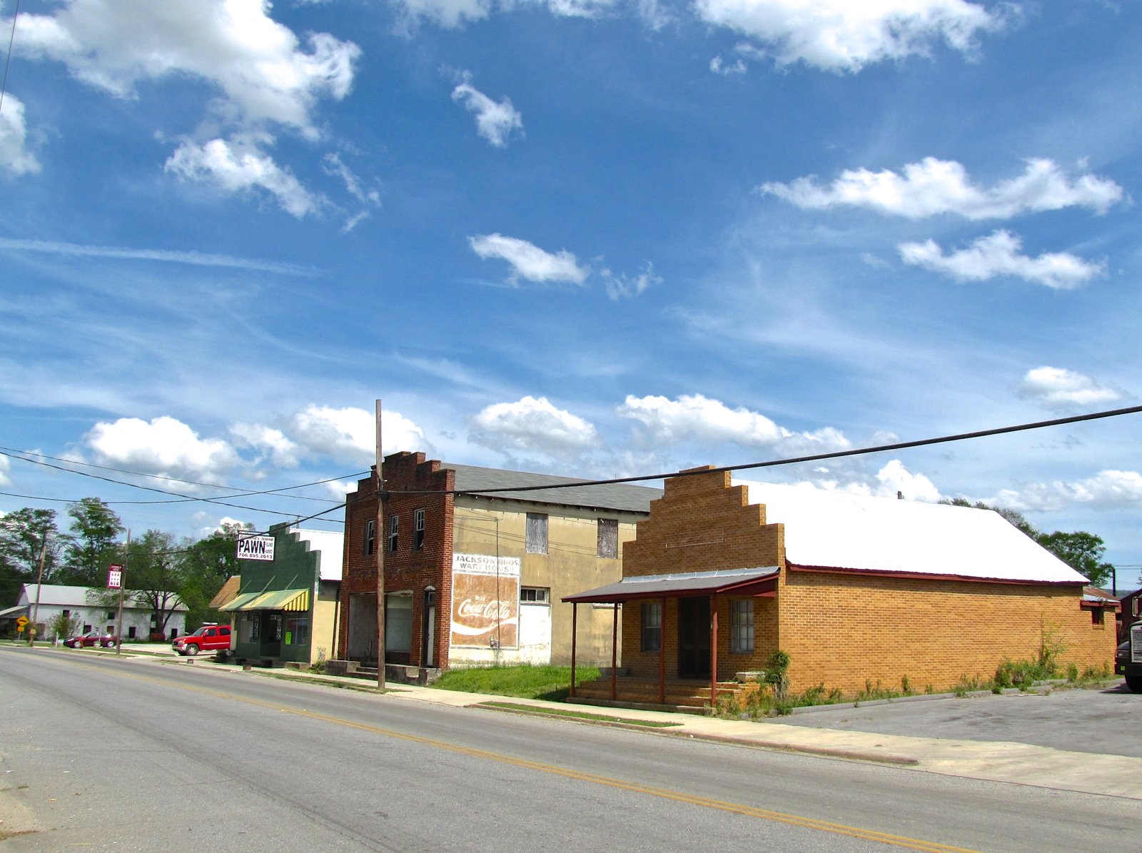 Old brick and stucco storefronts, one with a faded Coca-Cola mural, line a two-lane road under a blue sky with white clouds.