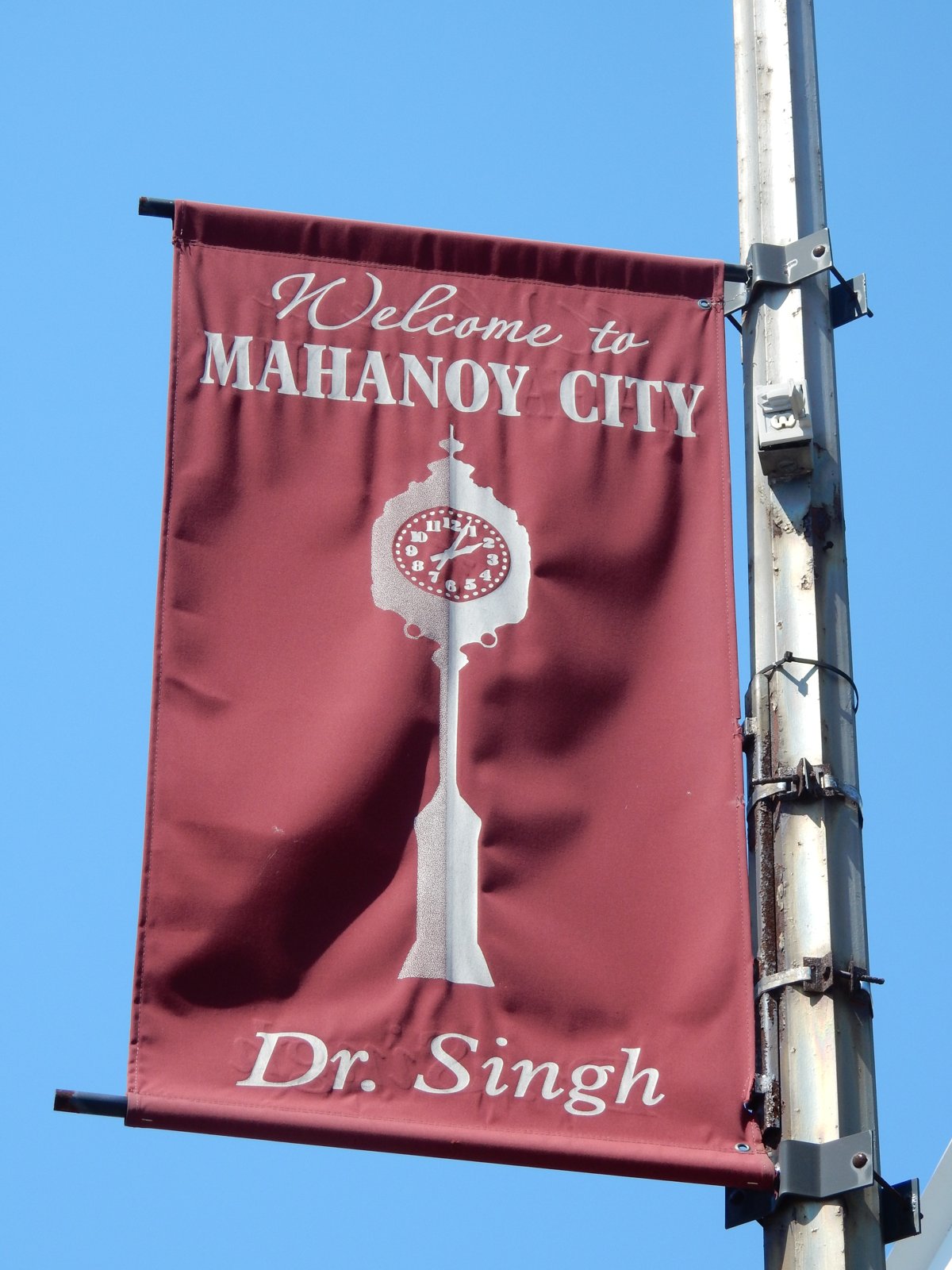 A maroon banner on a lamppost reading Welcome to Mahanoy City, with an illustration of a clock tower, against a blue sky.