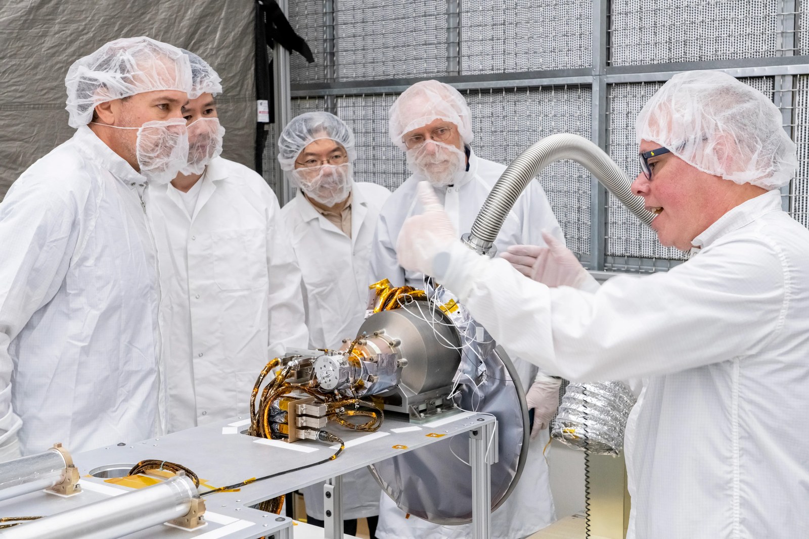 Several people in clean-room gowns gather around a cylindrical instrument on a workbench in a laboratory.
