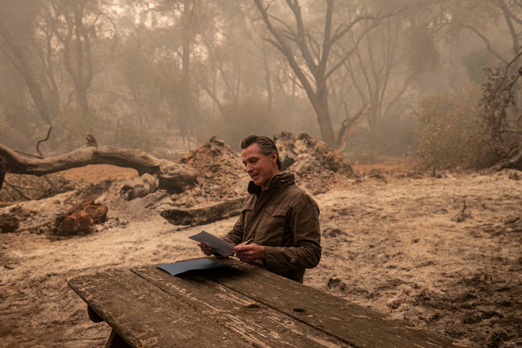 A man in a dark jacket sits at a weathered picnic table in a smoky, burned forest clearing, signing a document.