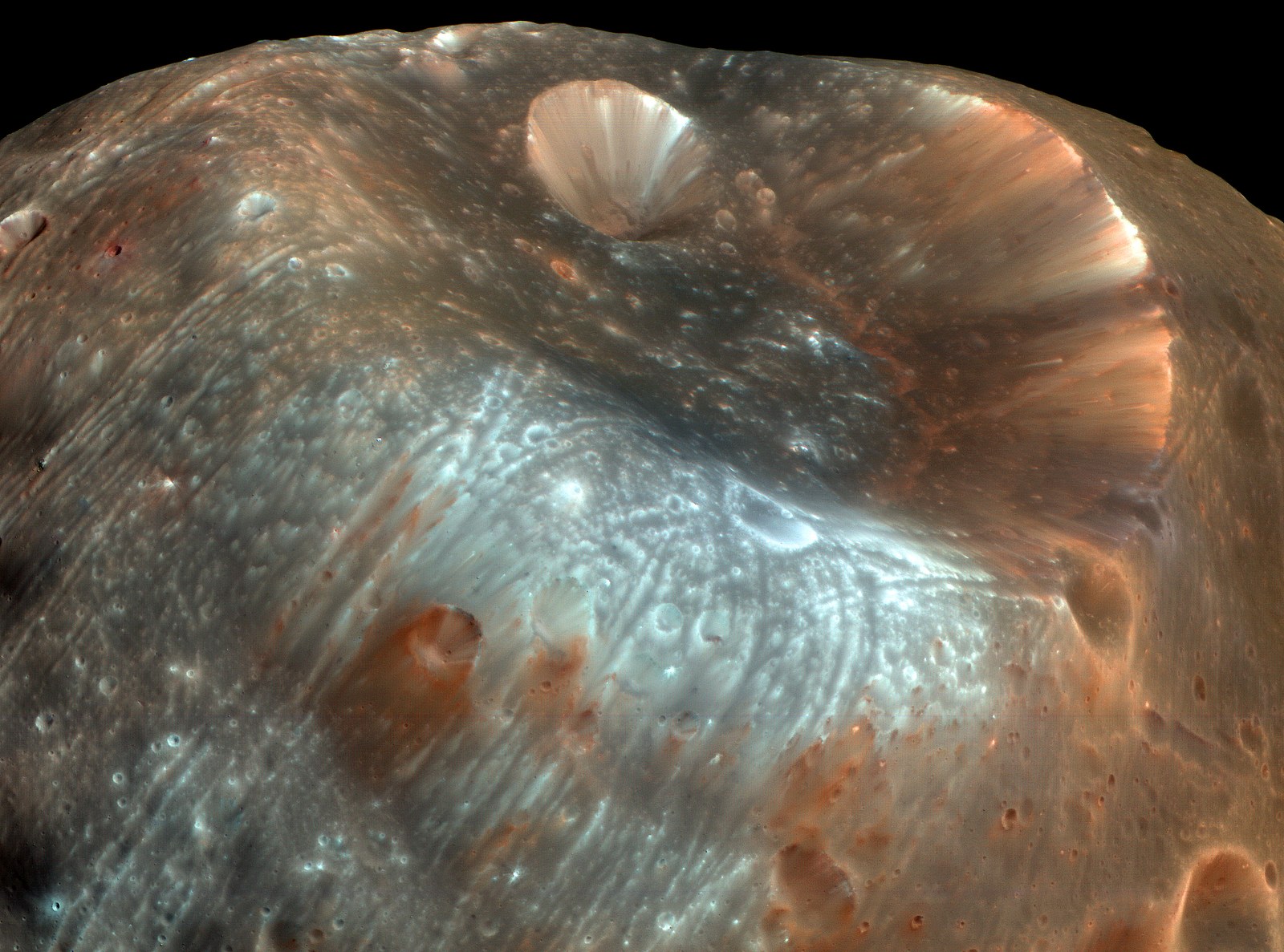 A close view of a wide, shallow crater on the gray surface of Phobos, with grooves and streaks running across the terrain.