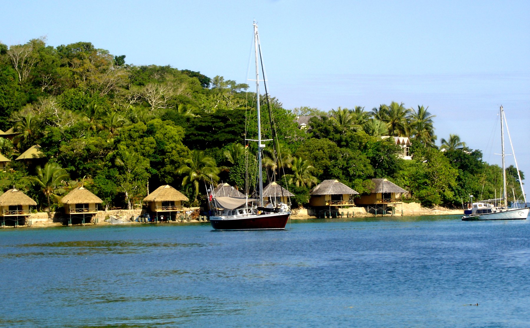 A sailboat anchored on blue water in Port Vila Harbour, Vanuatu, before a row of thatched waterfront bungalows and dense tropical trees.