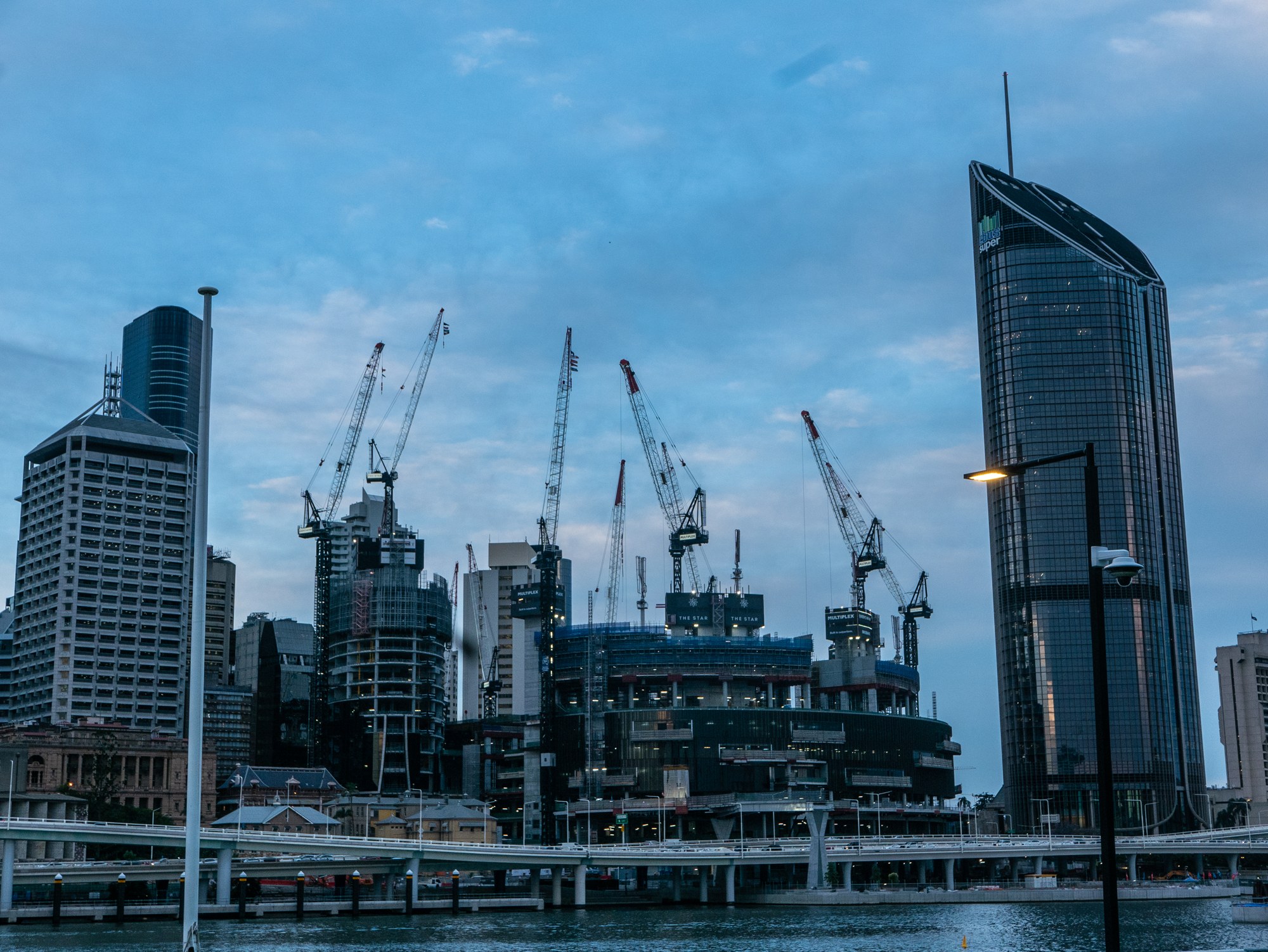 Construction cranes rising over the Queen's Wharf development on the Brisbane River at dusk, with city towers on either side and a riverside walkway in the foreground.