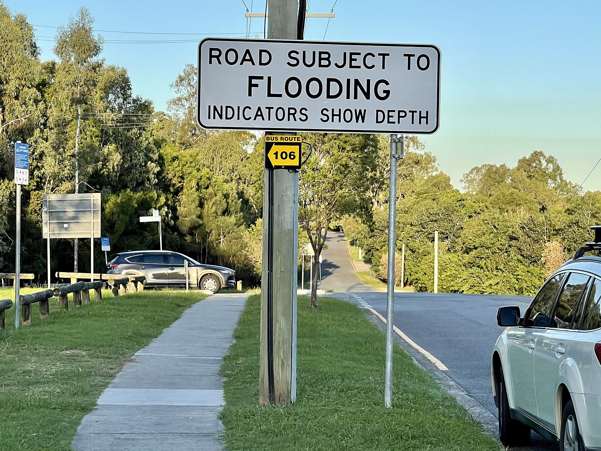 A white roadside sign reading Road Subject to Flooding, Indicators Show Depth, mounted on a pole beside a suburban street in Corinda, Queensland.