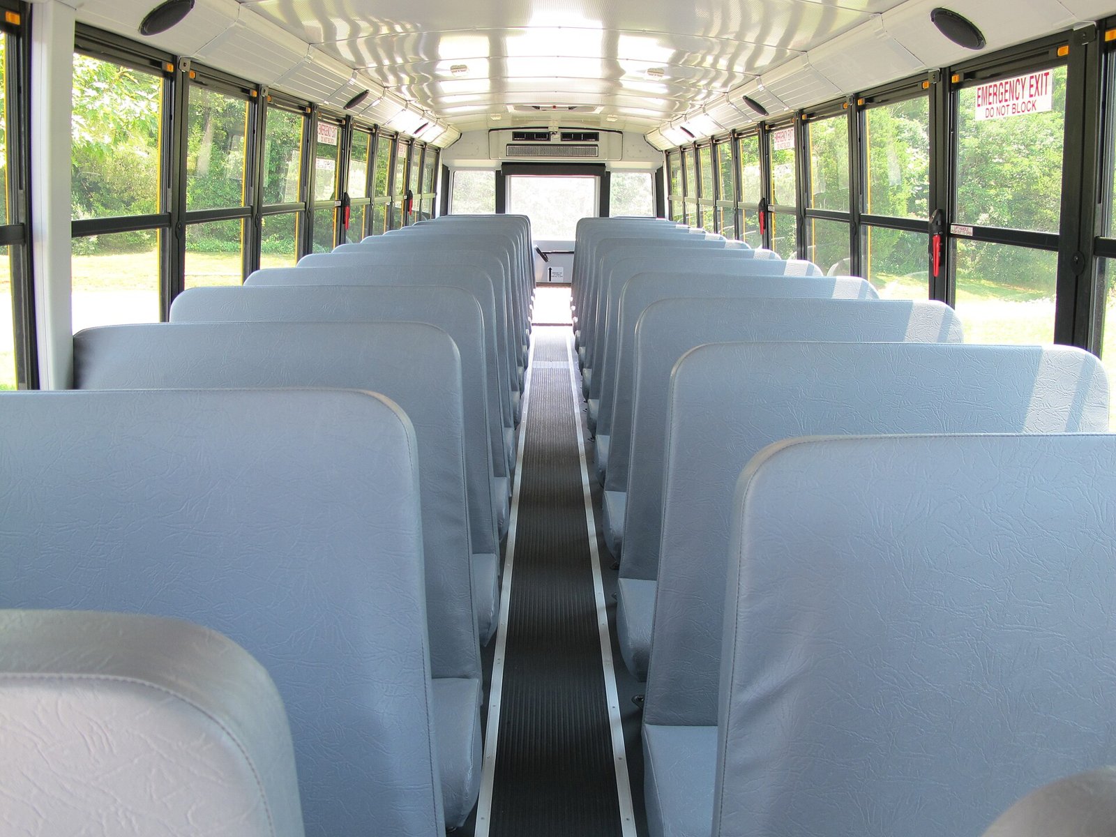 Rows of empty pale blue bench seats on either side of the aisle of a school bus, looking toward the rear emergency door.