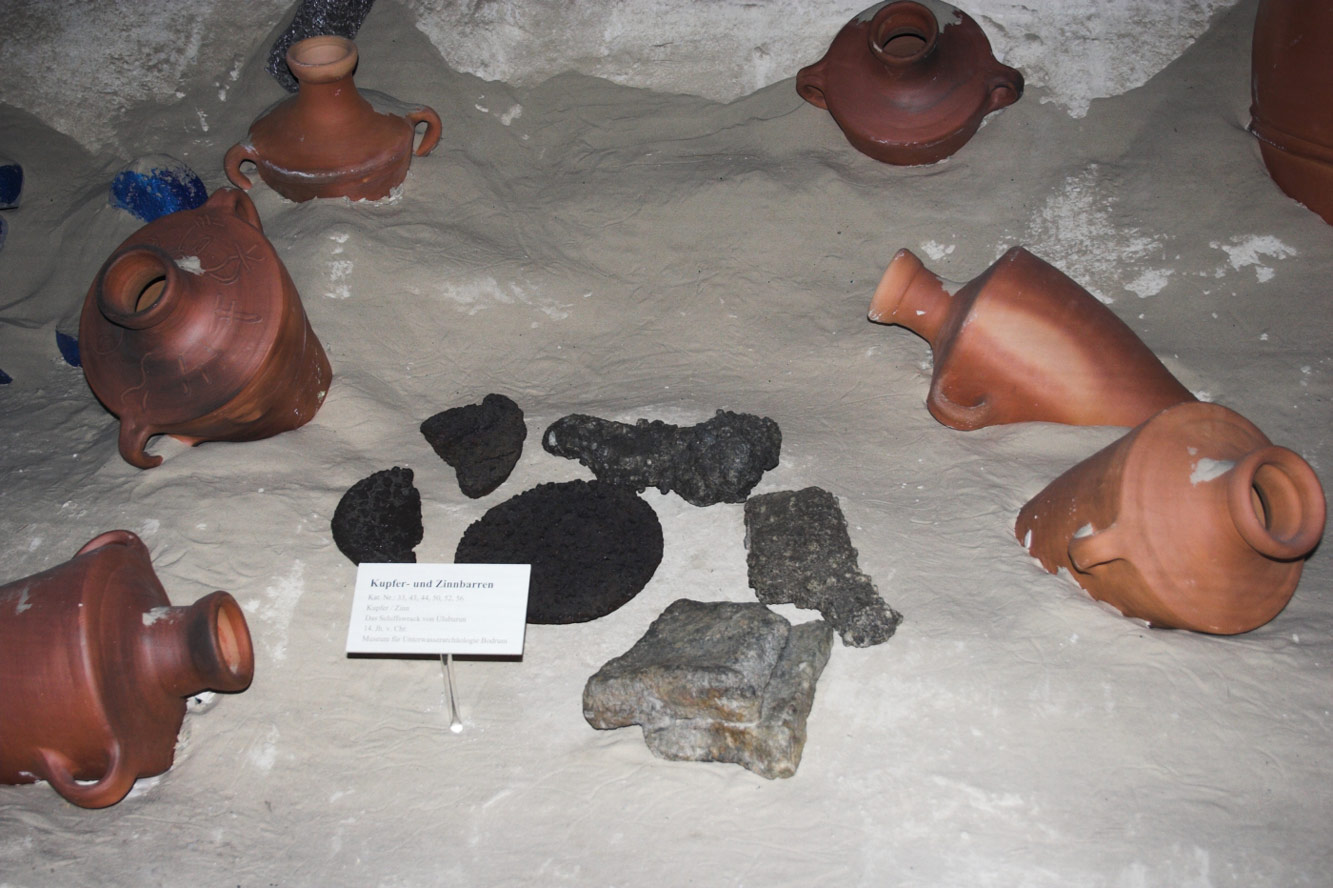 Museum display of ceramic jars and dark stone anchors laid out on pale sand to recreate a seabed wreck site.