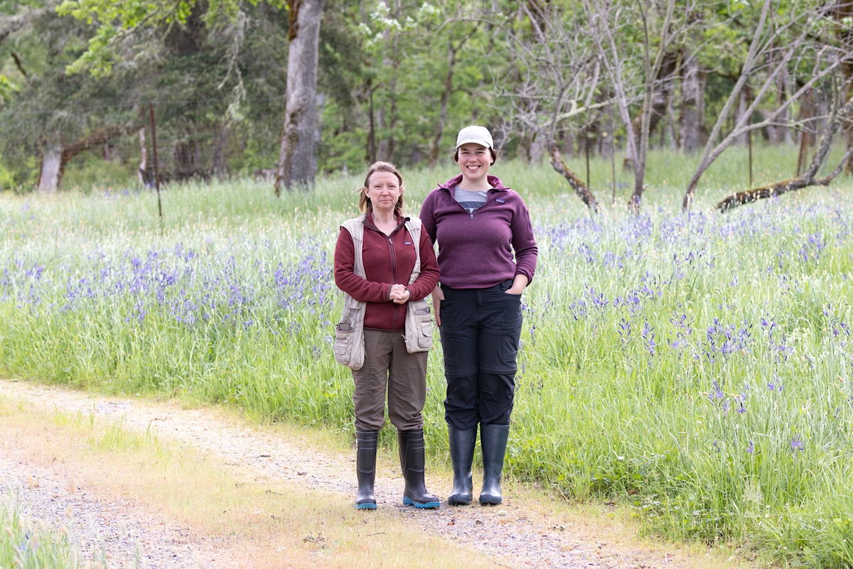 Two women in warm clothes and rubber boots stand beside a camas meadow.