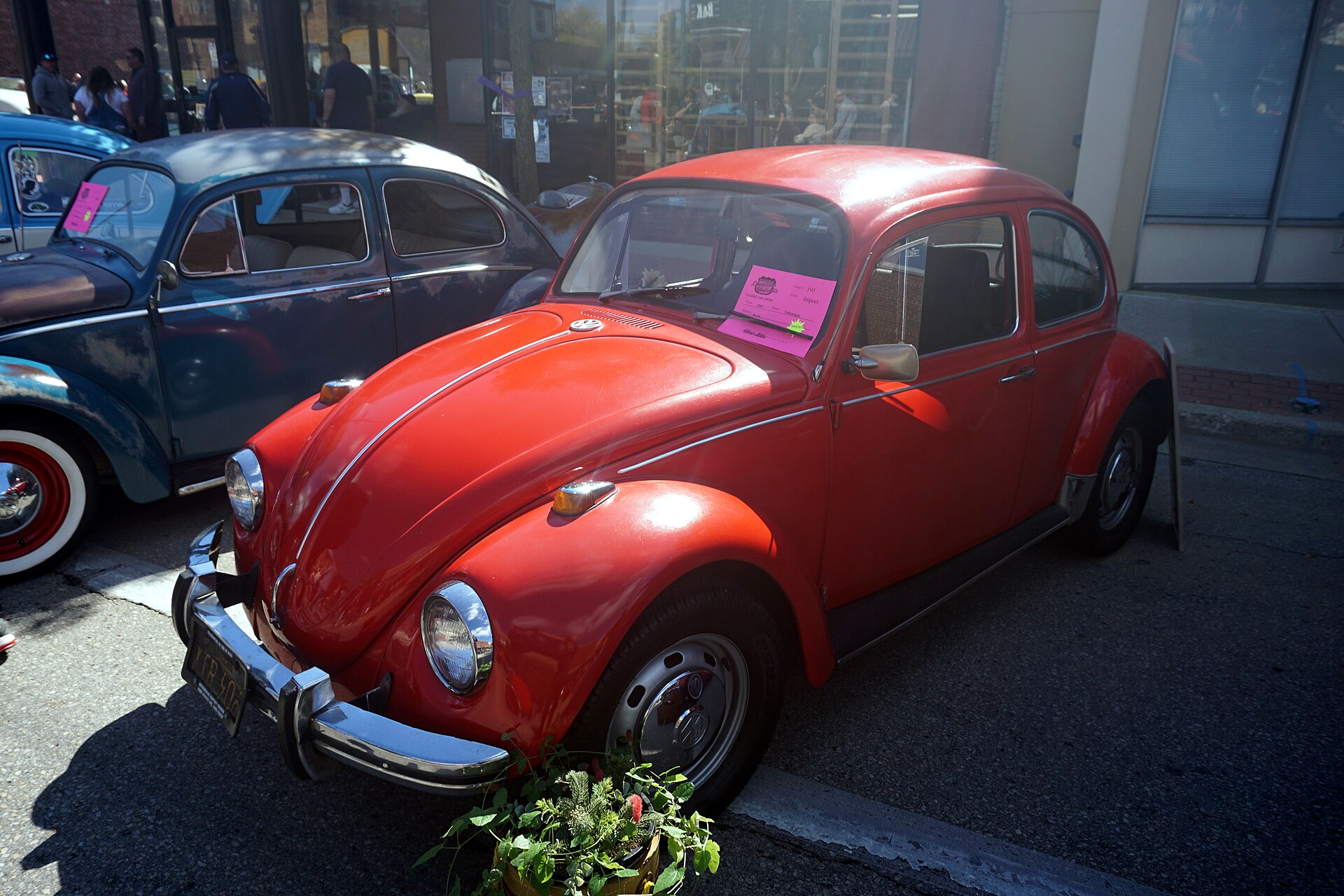 A red 1969 Volkswagen Beetle parked at a curb beside a dark blue Beetle at an outdoor car show.