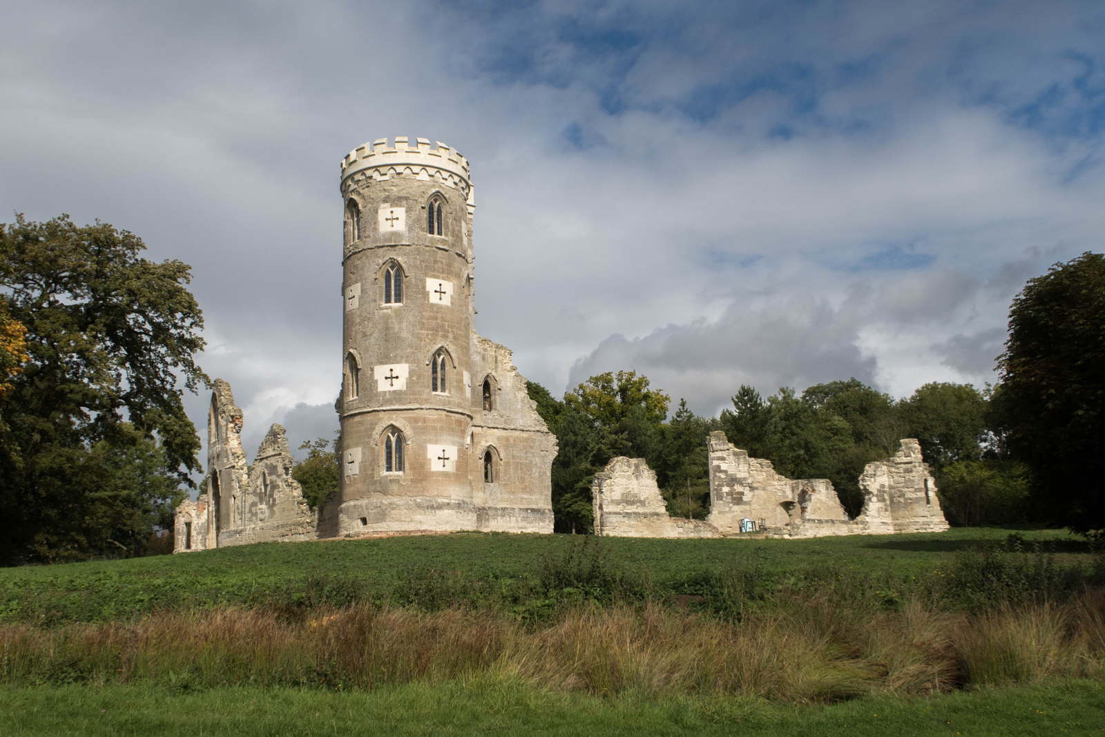 An 18th-century Gothic folly, a crenellated stone tower with ruined walls, on a grassy rise beneath trees and a cloudy sky.