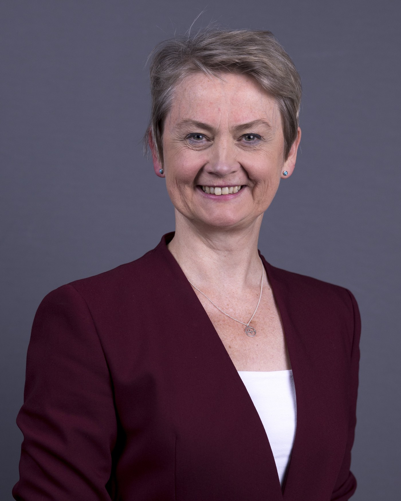 A smiling woman with short gray hair, in a burgundy jacket, in a formal portrait against a plain gray background.